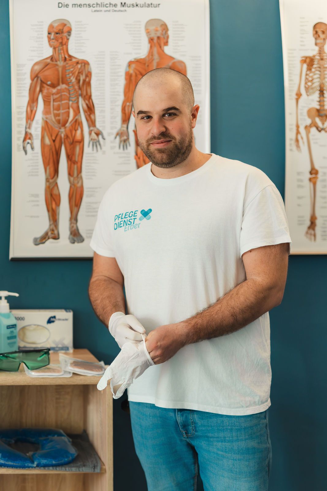 Ein Foto von DGKP Christian Erber vor einer blauen Wand. Auf dem T-Shirt ist das Logo von Pflegedienst Erber